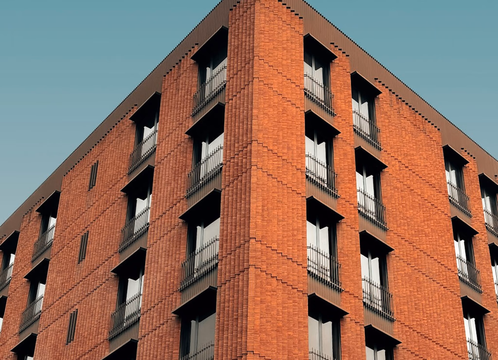 Modern red brick apartment or office building viewed from below against a blue sky, showcasing its geometric architecture with evenly spaced windows and balconies.
